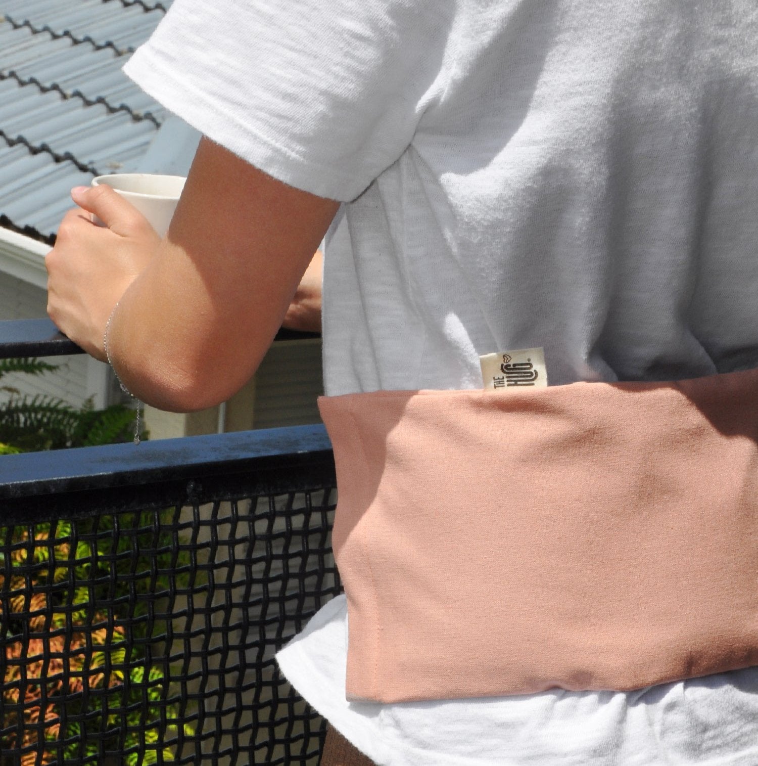 Person standing on balcony having a cup of tea and wearing The Hug across the back for back pain. The Hug featured has a blush colored cover.