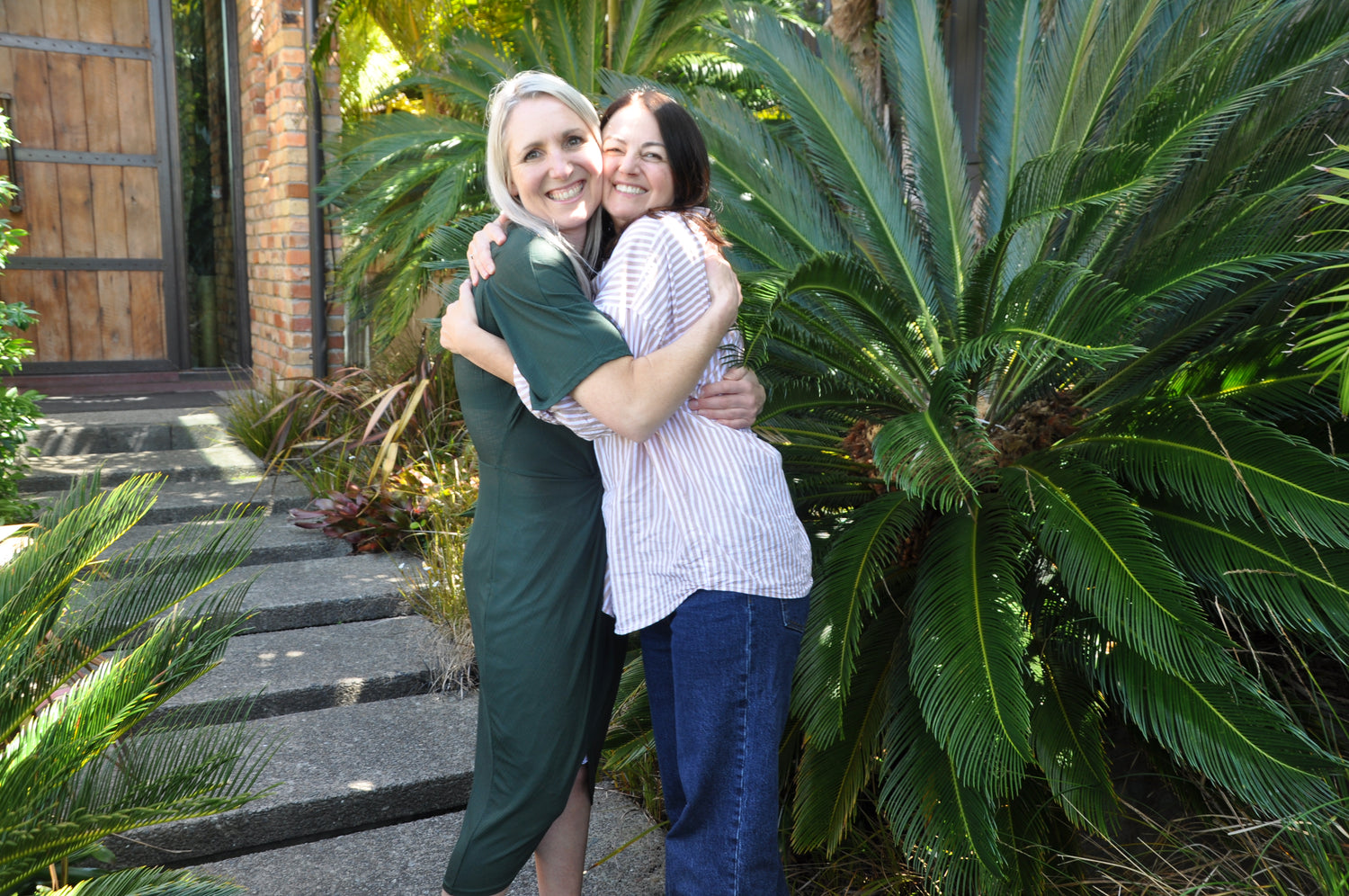 Nat and Nic founders of The Hug celebrate their collaborative success with a Hug. They are outside in  garden with a large Queen Palm beside them. The sun is shining and they are clearly very happy.