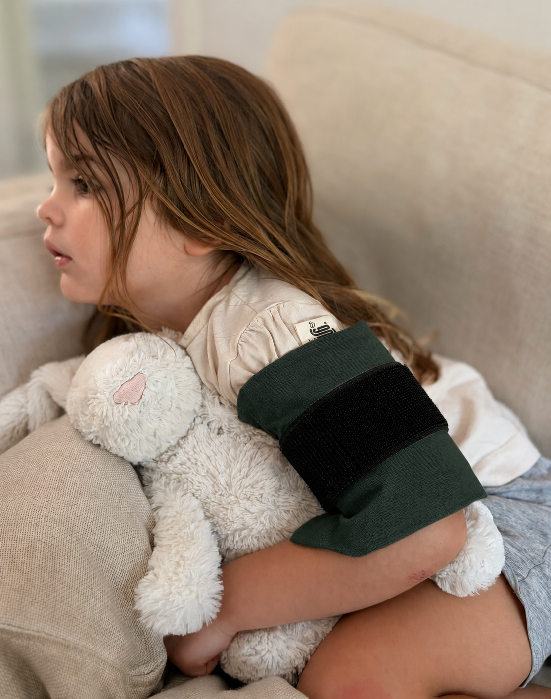 A young girl with long brown hair sits on a beige couch, hugging a white plush bunny. She is wearing a pine mini hug aon her upper arm.